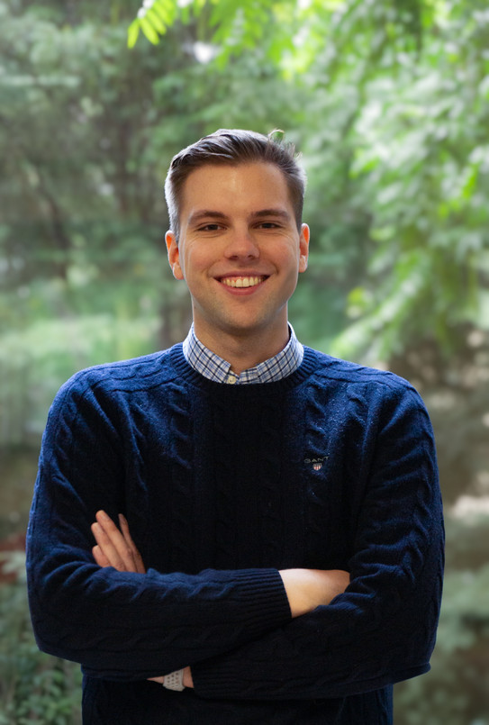 Ein Mann im dunkelblauen Pullover mit verschränkten Armen, einem starken Lächeln und im Hintergrund eine grüne Landschaft