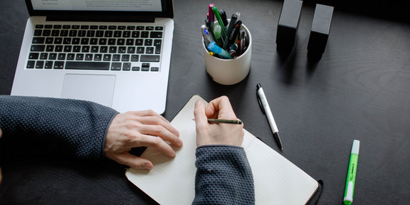 A student working at a table with a laptop, pencils and a notebook.