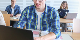 Student sitzt im Seminarraum am Laptop, im Hintergrund weitere Studierende